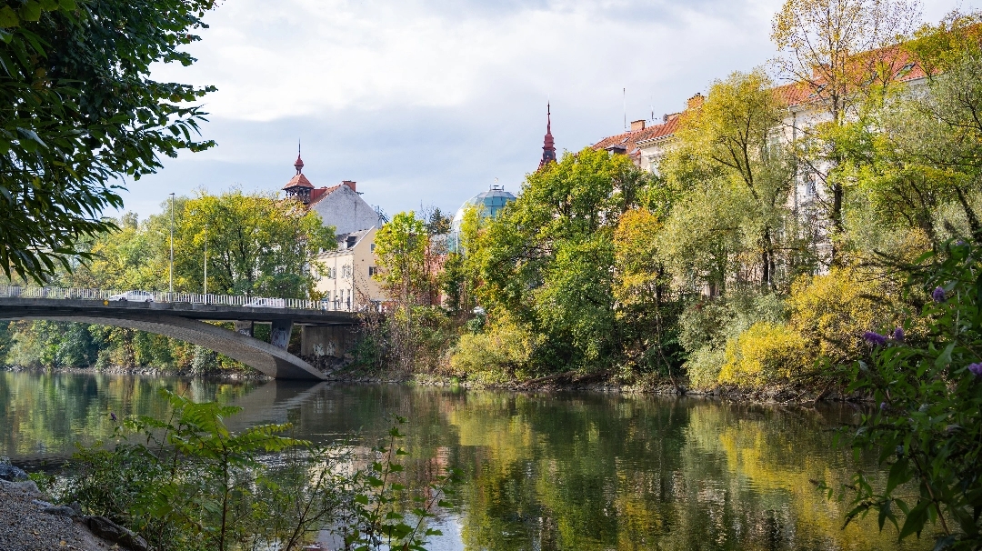 Blick auf eine Brücke über einen ruhigen Fluss, umgeben von Bäumen in herbstlichen Farben und historischen Gebäuden im Hintergrund.