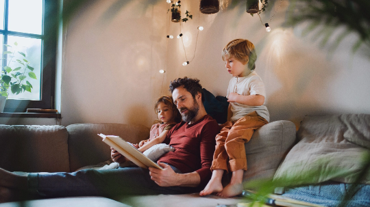 Ein Mann sitzt mit zwei kleinen Kindern auf einem Sofa in einem gemütlich beleuchteten Wohnzimmer. Er hält ein aufgeschlagenes Buch in der Hand und liest daraus vor. Ein Kind sitzt neben ihm, das andere lehnt sich von der Sofalehne herüber, um mit ins Buch zu schauen. Im Hintergrund hängen kleine Pflanzen und Lichterketten an der Wand, und durch ein Fenster fällt Tageslicht herein.