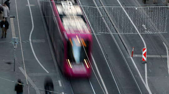 Eine pinkfarbene Straßenbahn fährt schnell auf Schienen durch eine Stadtstraße. Das Bild ist leicht verwischt, was die Bewegung der Bahn betont. Auf beiden Seiten der Gleise sind Fußgänger zu sehen, und über der Straße hängen Lichterketten.