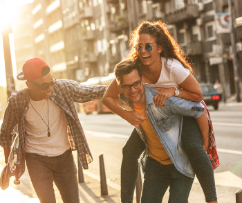 Drei junge Menschen haben Spaß auf einer sonnigen Stadtstraße. Eine Frau mit Sonnenbrille lacht und wird von einem Mann auf dem Rücken getragen, während ein dritter Freund daneben läuft und ein Skateboard in der Hand hält. Alle drei wirken fröhlich und ausgelassen, das Sonnenlicht fällt warm auf die Szene. Im Hintergrund sind Wohnhäuser und parkende Autos zu sehen.