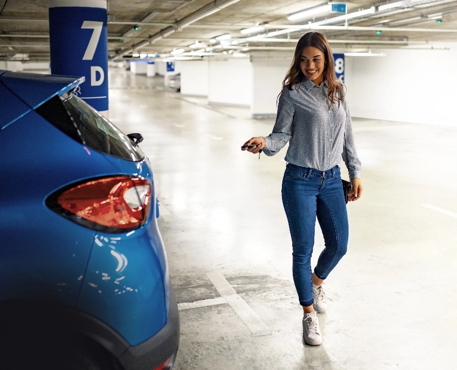 Eine junge Frau steht in einem hell beleuchteten Parkhaus neben einem blauen Auto. Sie trägt eine hellgraue Bluse, enge Jeans und weiße Turnschuhe. In der rechten Hand hält sie einen Autoschlüssel und schaut lächelnd auf das Auto, als würde sie es gerade auf- oder zuschließen. Im Hintergrund sind blau-weiße Säulen mit den Beschriftungen „7D“ und „8D“ zu sehen.