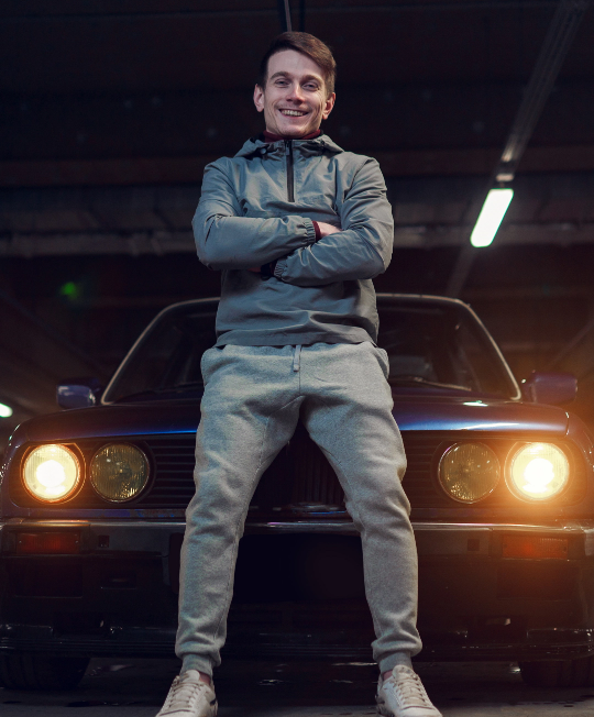 Ein junger Mann sitzt auf der Motorhaube eines dunklen Autos in einer Tiefgarage. Er trägt eine graue Trainingsjacke, graue Jogginghose und weiße Turnschuhe. Die Arme sind vor der Brust verschränkt, und er lächelt in die Kamera. Die Scheinwerfer des Autos sind eingeschaltet und leuchten warmgelb. Die Szene ist leicht gedimmt beleuchtet.
