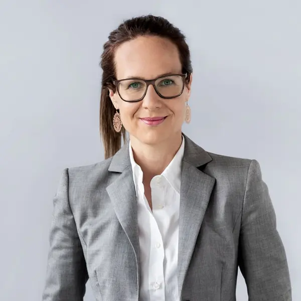 Eine Person mit Brille und Ohrringen trägt eine graue Anzugjacke über einem weißen Hemd. Der Hintergrund ist einfarbig hellgrau.