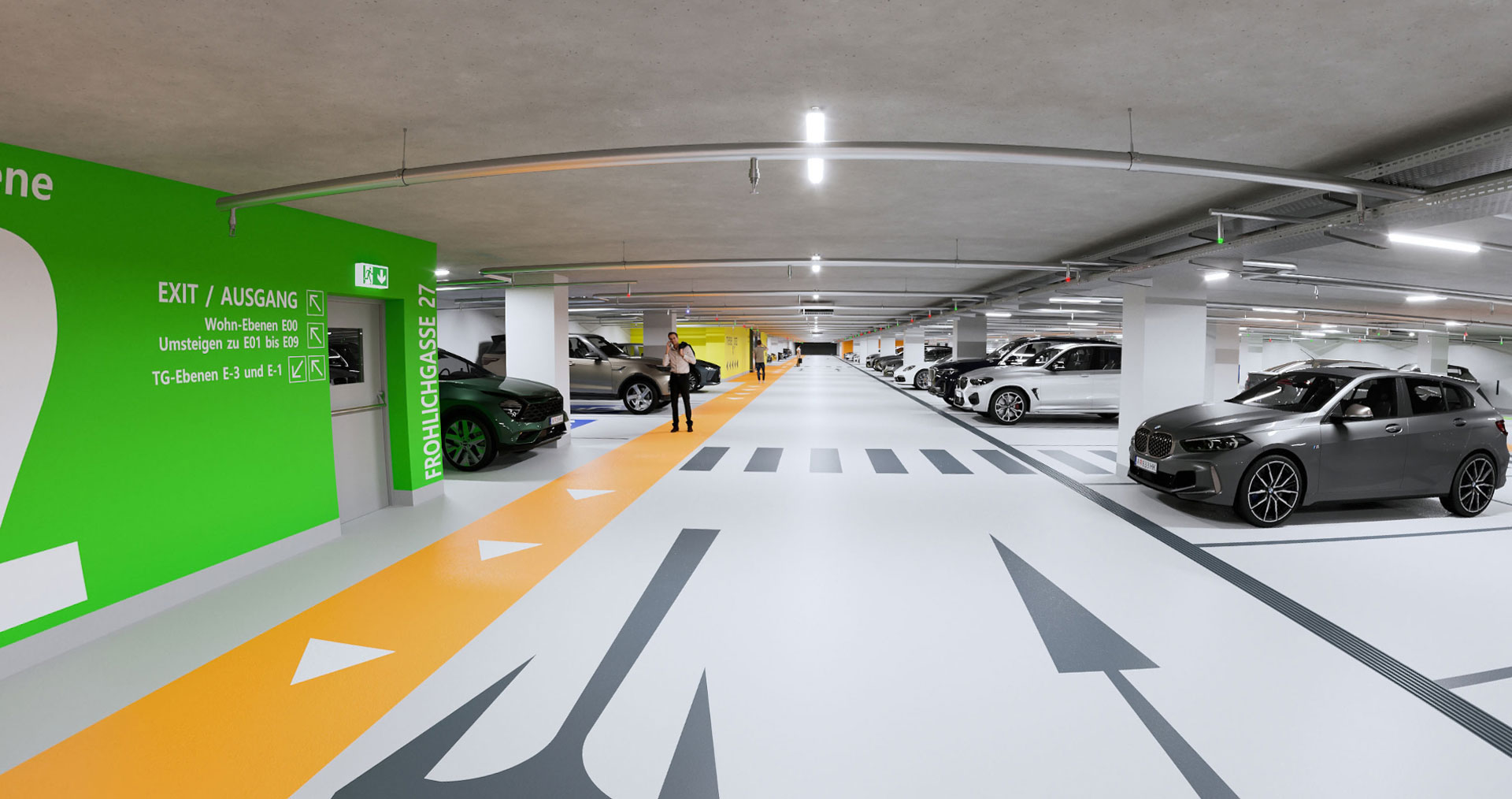 Helle Tiefgarage mit Autos und ein Mann mit Telefon, links ein grüner Aufgang zum Wohngebäude